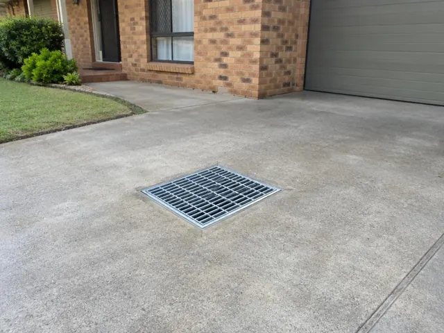 Concrete driveway with a clean metal drain grate beside a brick house and garage door in Smithfield